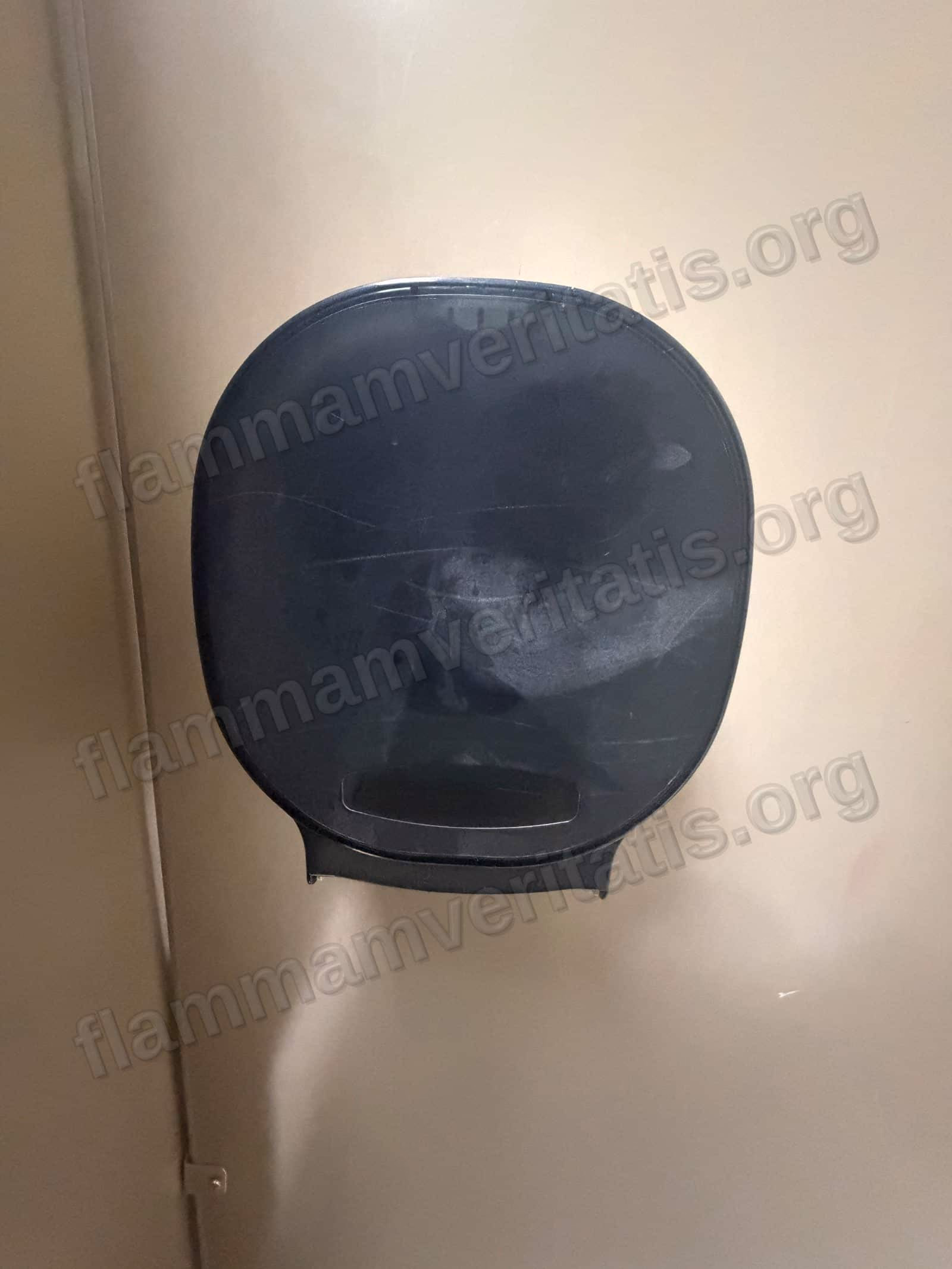 Despachador de papel higiénico de plástico negro montado en la pared del cubículo, rayado y sin papel a la vista.