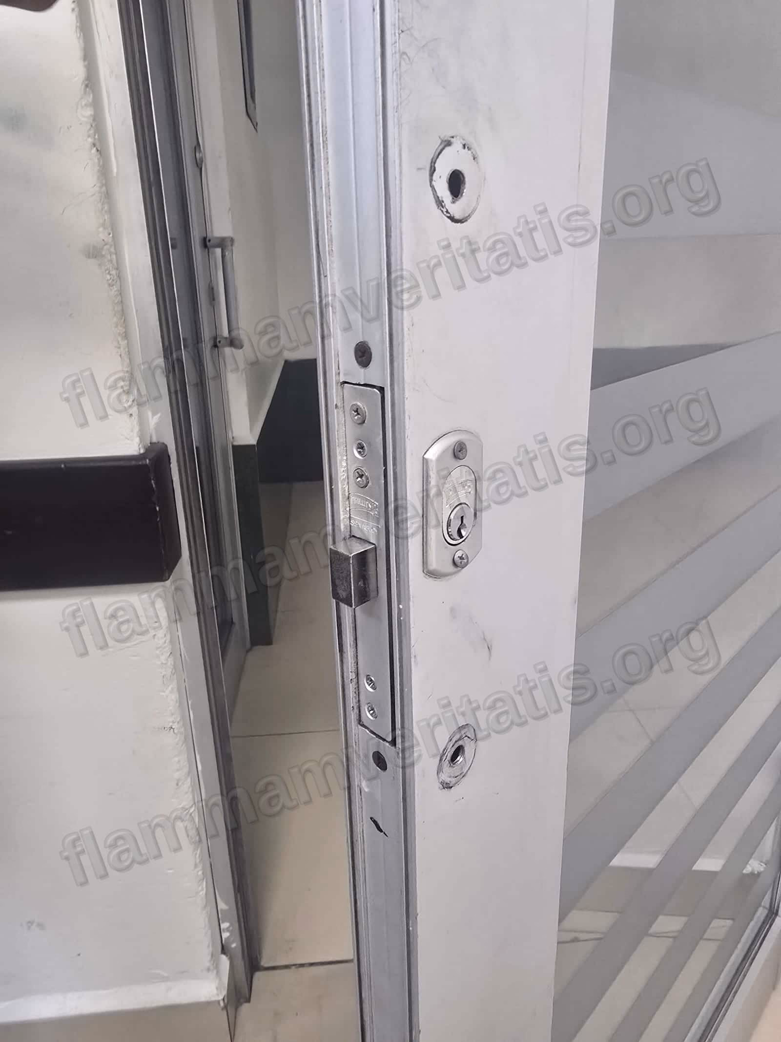 Canto de una puerta corrediza de vidrio y aluminio en el aula 2107, edificio 2, con una chapa de seguridad y dos orificios de montaje vacíos, sin manija instalada.