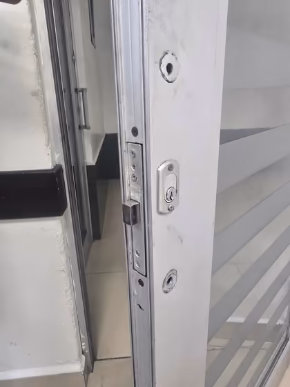 Canto de una puerta corrediza de vidrio y aluminio en el aula 2107, edificio 2, con una chapa de seguridad y dos orificios de montaje vacíos, sin manija instalada.
