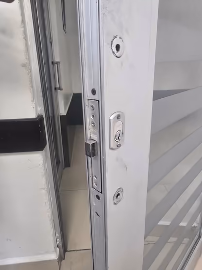 Canto de una puerta corrediza de vidrio y aluminio en el aula 2107, edificio 2, con una chapa de seguridad y dos orificios de montaje vacíos, sin manija instalada.