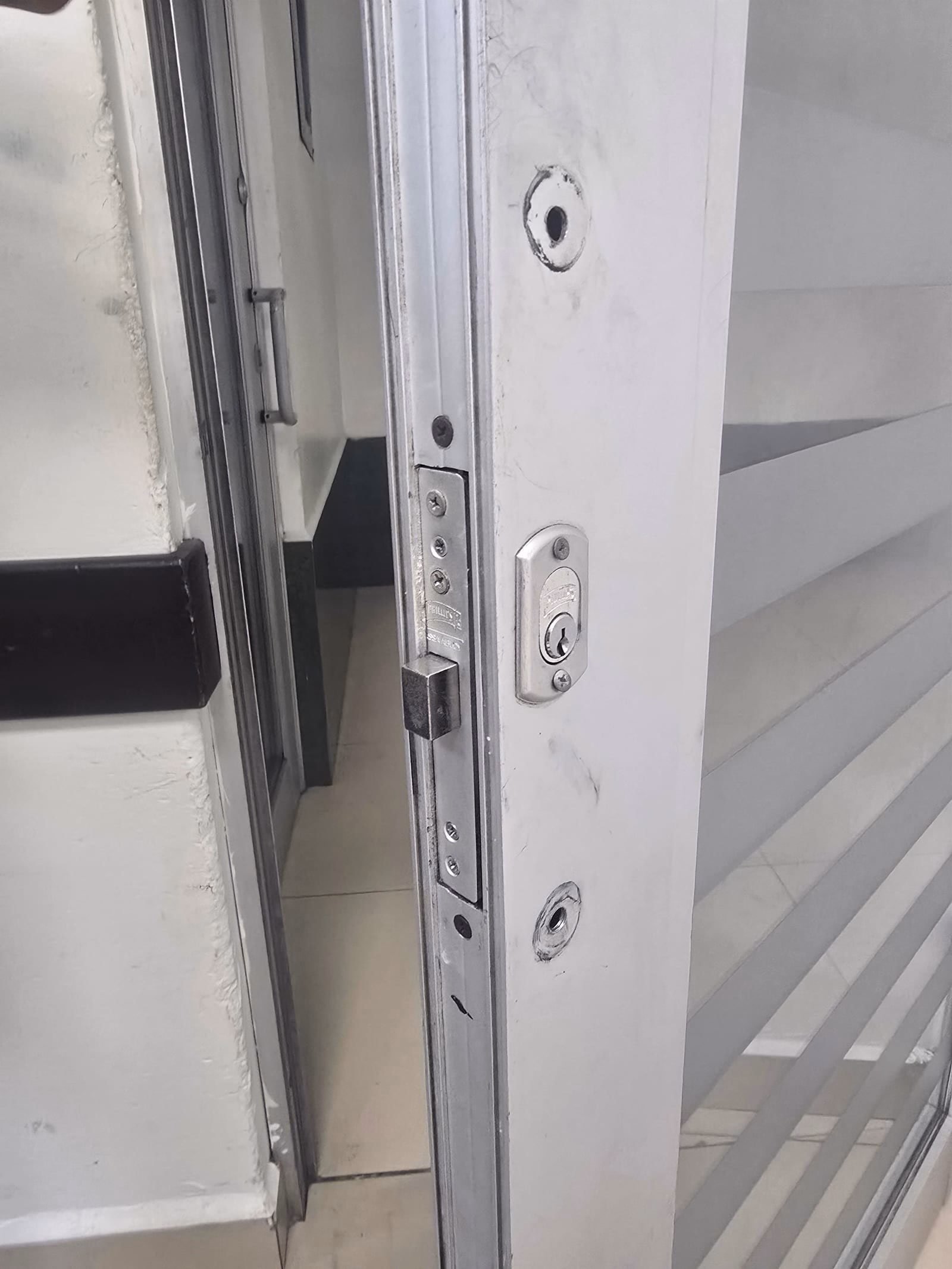 Canto de una puerta corrediza de vidrio y aluminio en el aula 2107, edificio 2, con una chapa de seguridad y dos orificios de montaje vacíos, sin manija instalada.